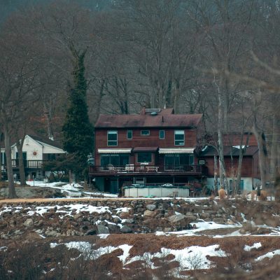 Tranquil lakeside house surrounded by snow patches and bare trees, highlighting the serene winter landscape.